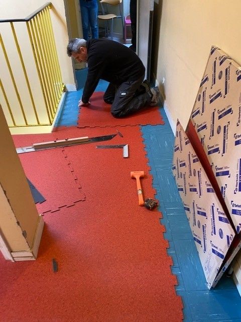 Un ouvrier agenouillé sur le sol rouge d'un couloir, près d'une bordure bleue, utilise des outils à côté d'un panneau emballé.