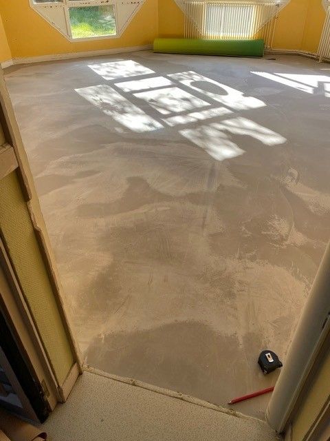 Pièce vide avec sol en béton brut, lumière du soleil entrant par les fenêtres, et une serpillière et une pelle près de la porte.