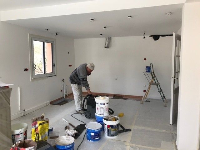 Un ouvrier ponce un sol dans une pièce blanche à l'aide de seaux de peinture et d'une échelle lors de travaux de rénovation.