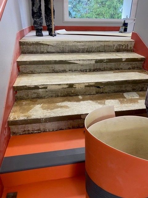 Escalier intérieur avec plusieurs marches en béton usées, sol orange et un grand tube en carton au premier plan.