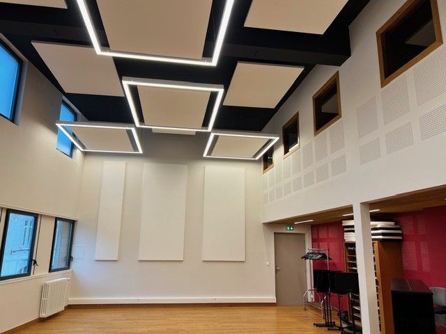 Salle de musique moderne vide avec panneaux acoustiques au plafond, murs blancs, fenêtres et rideaux rouges.