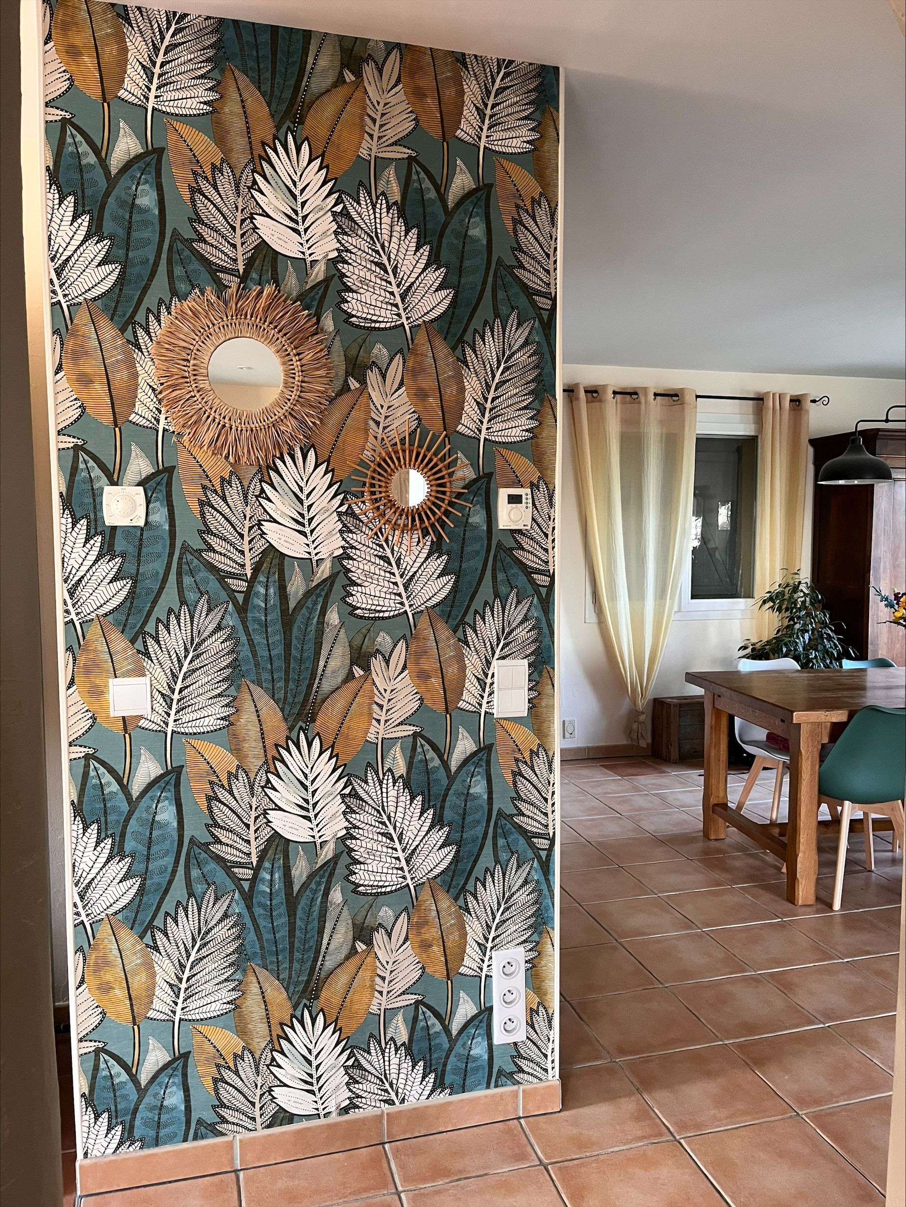 Un mur d'accent orné de papier peint à motifs floraux mène à une salle à manger lumineuse avec une table en bois et des rideaux.