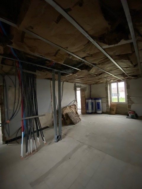 Pièce inachevée avec poutres apparentes au plafond, fils électriques pendants et matériaux de construction près d'une fenêtre.