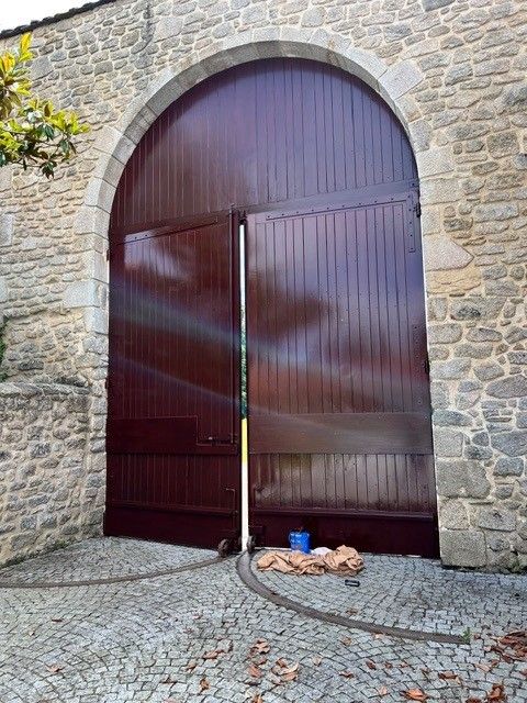 Grande porte cintrée en bois encastrée dans un mur de pierre, avec un seau bleu et une corde posés sur des pavés en contrebas.