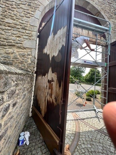Un grand panneau de portail sombre est installé à côté d'un mur de pierre, avec un ouvrier sur un échafaudage en arrière-plan.
