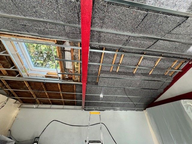 Plafond en construction avec isolation apparente, poutres de soutien rouges et ouverture de puits de lumière