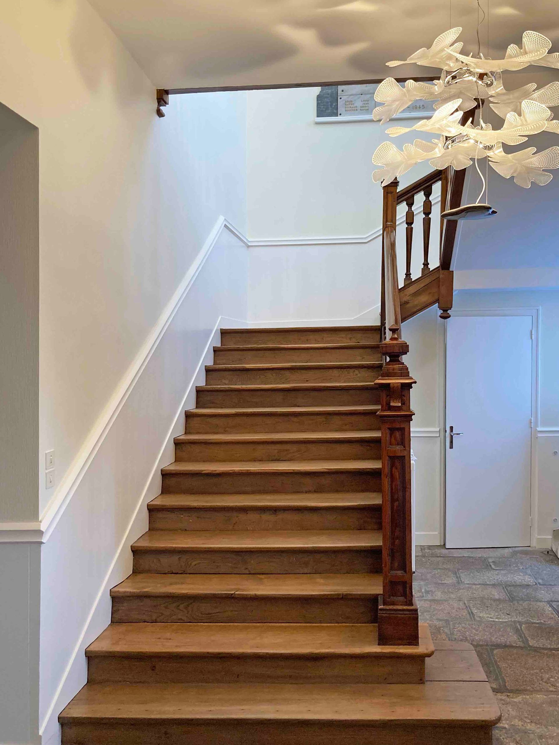 Escalier aux marches en bois et aux murs blancs, menant à un palier supérieur orné d'une balustrade et surmonté d'un lustre.