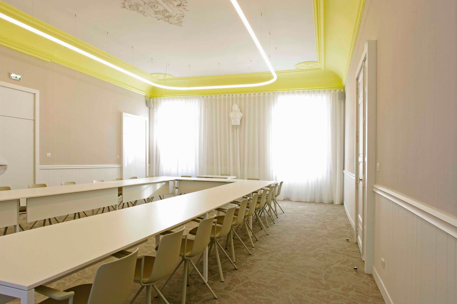 Salle de conférence vide avec de longues tables, des chaises et de grandes fenêtres éclairées par des plafonniers jaunes.