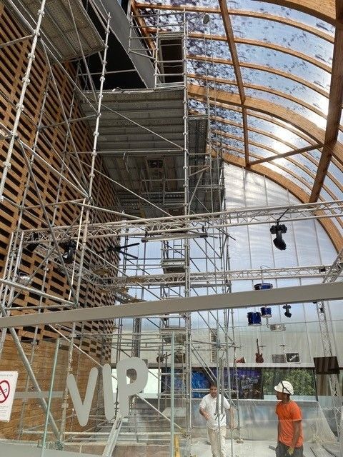 Haut atrium vitré en construction avec échafaudages, un ouvrier portant un casque orange et de grandes lettres.