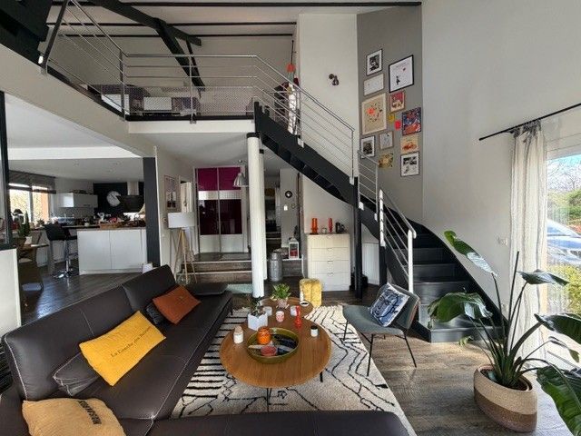 Salon moderne avec canapé noir, escalier, table basse et grandes fenêtres