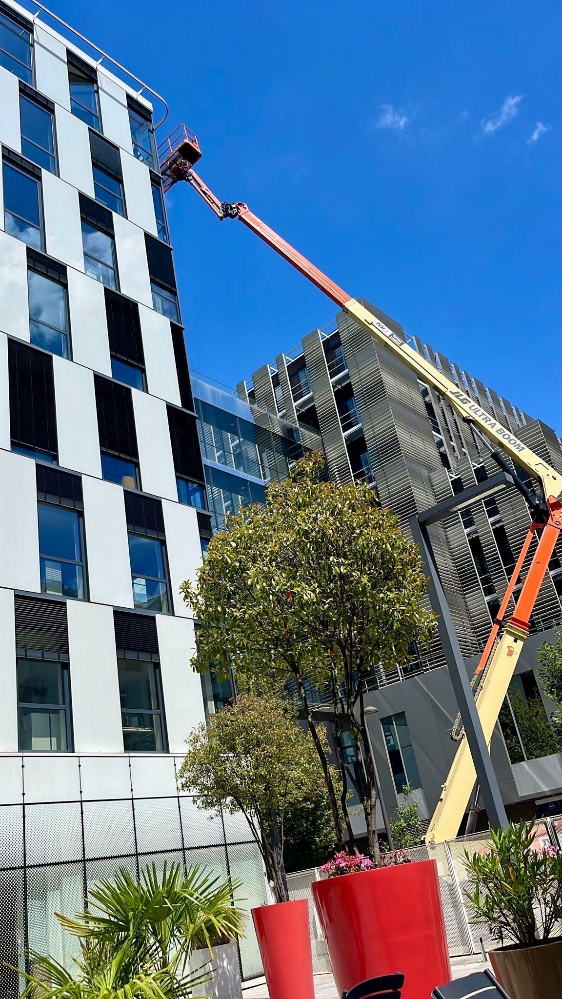 Une grue soulève un grand panneau de fenêtre près d'un bâtiment, maintenu par un dispositif de ventouse rouge.