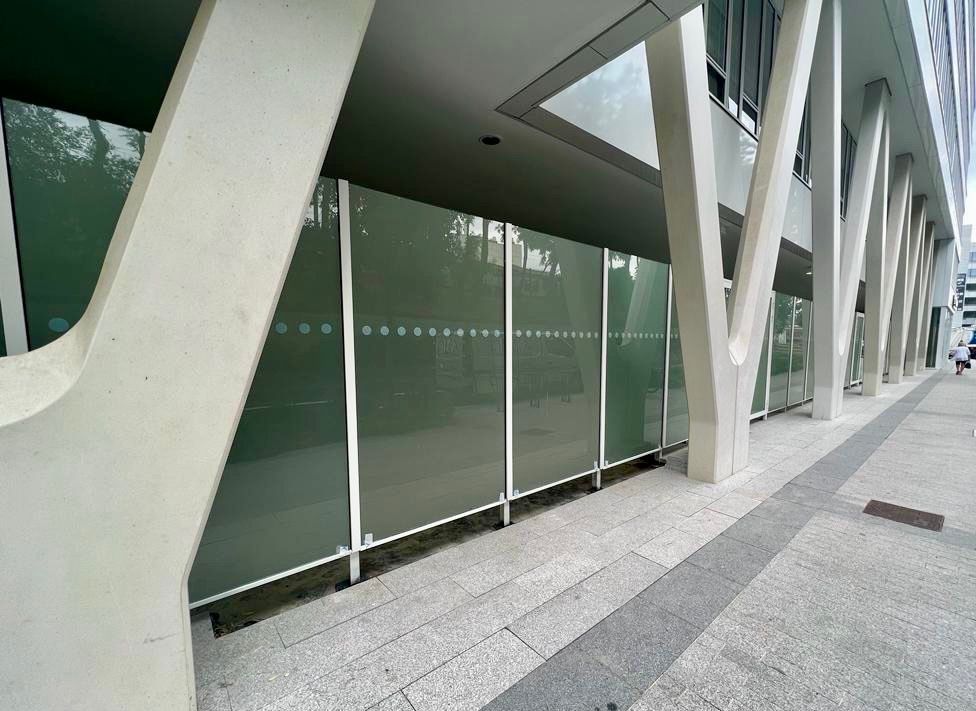 Façade d'un bâtiment moderne avec poutres de soutien blanches, panneaux de verre dépoli et trottoir.