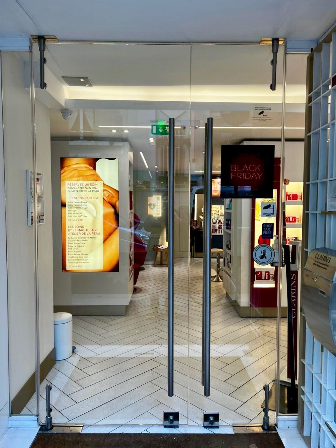 Des doubles portes vitrées donnent sur un magasin bien éclairé, avec des présentoirs et une personne à l'intérieur.