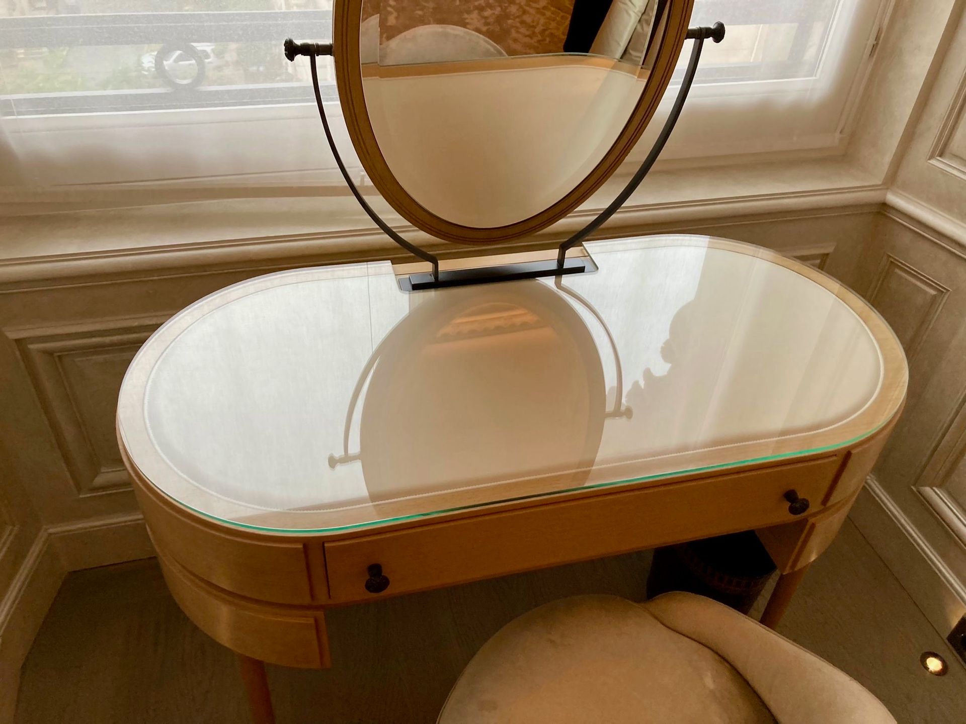 Coiffeuse avec miroir ovale, plateau en verre et tabouret rembourré couleur crème devant une fenêtre.