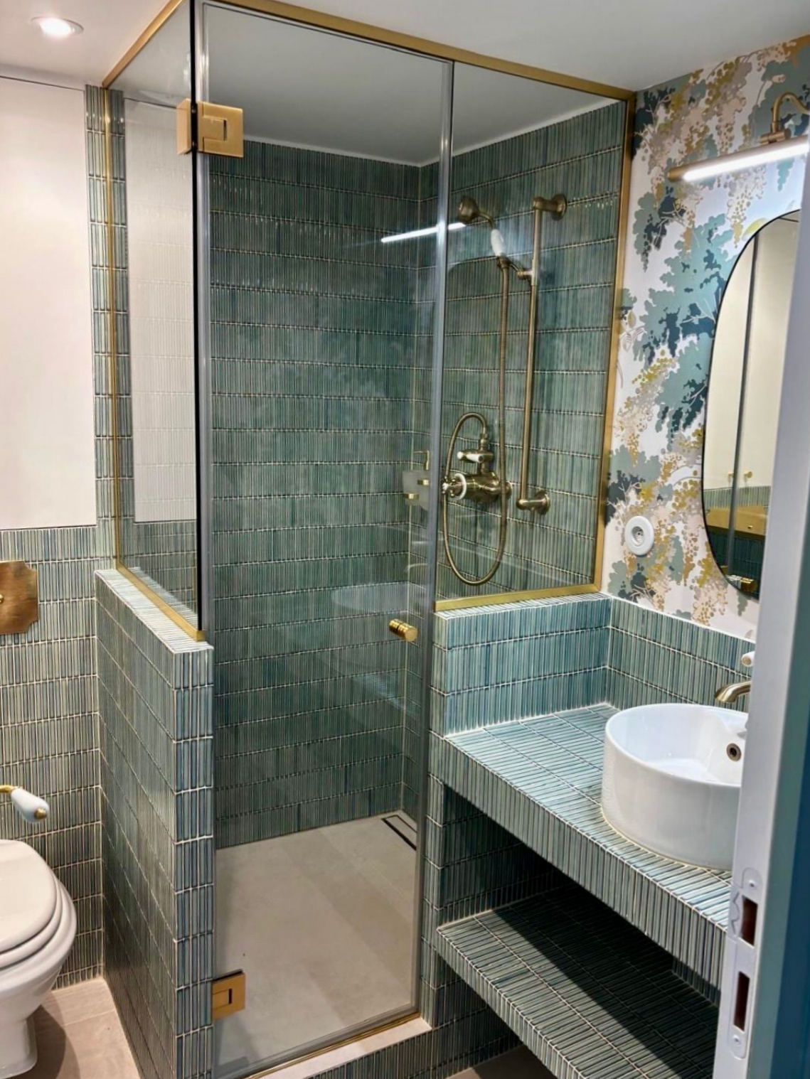 Salle de bains carrelée de vert avec douche en verre, robinetterie dorée et papier peint à motifs. Lavabo et toilettes blancs.