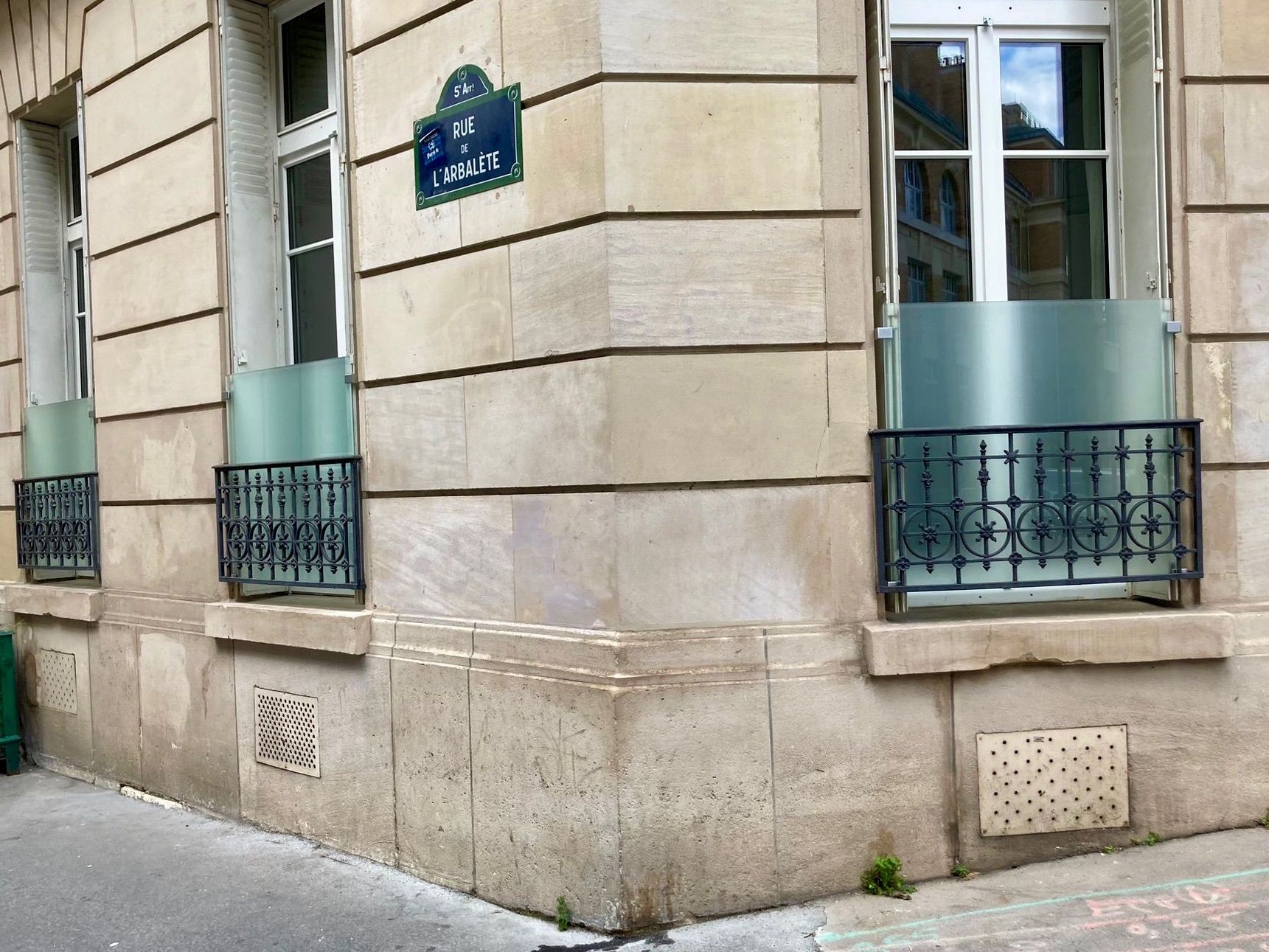 Coin d'un bâtiment en pierre beige avec fenêtres, grilles en fer forgé et panneau de verre.