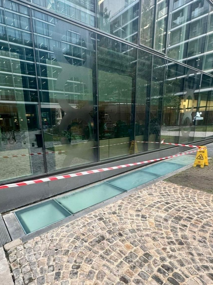 Façade d'un bâtiment aux parois de verre, avec un panneau de signalisation et du ruban adhésif rouge et blanc marquant une surface vitrée au niveau du sol.