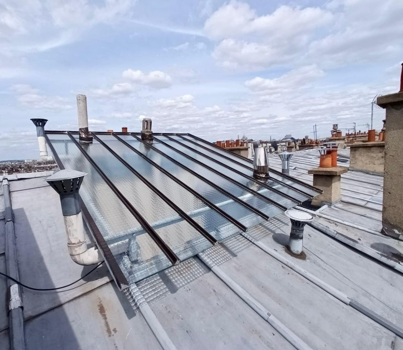 Un toit avec des panneaux de verre, des cheminées et des aérations sous un ciel partiellement nuageux.