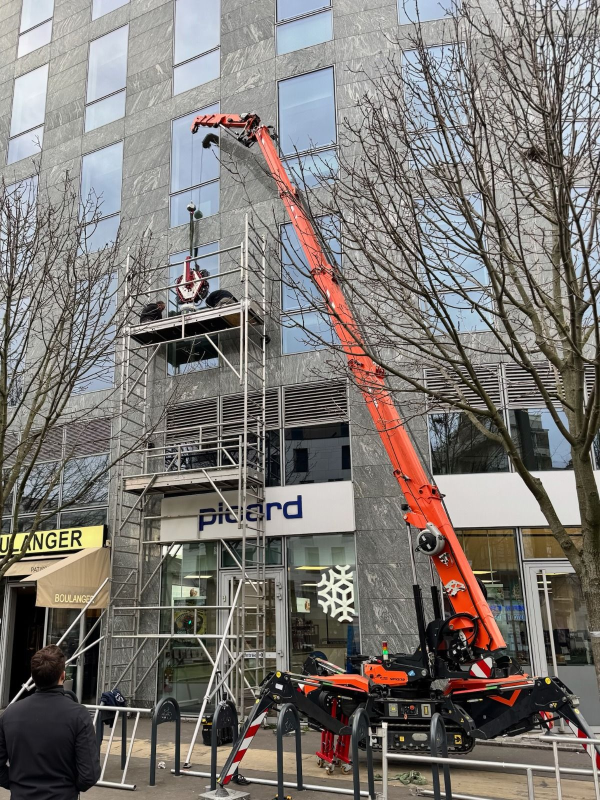 Une grande grue orange s'étend vers un bâtiment. Des échafaudages se trouvent près de l'entrée d'un magasin Picard.