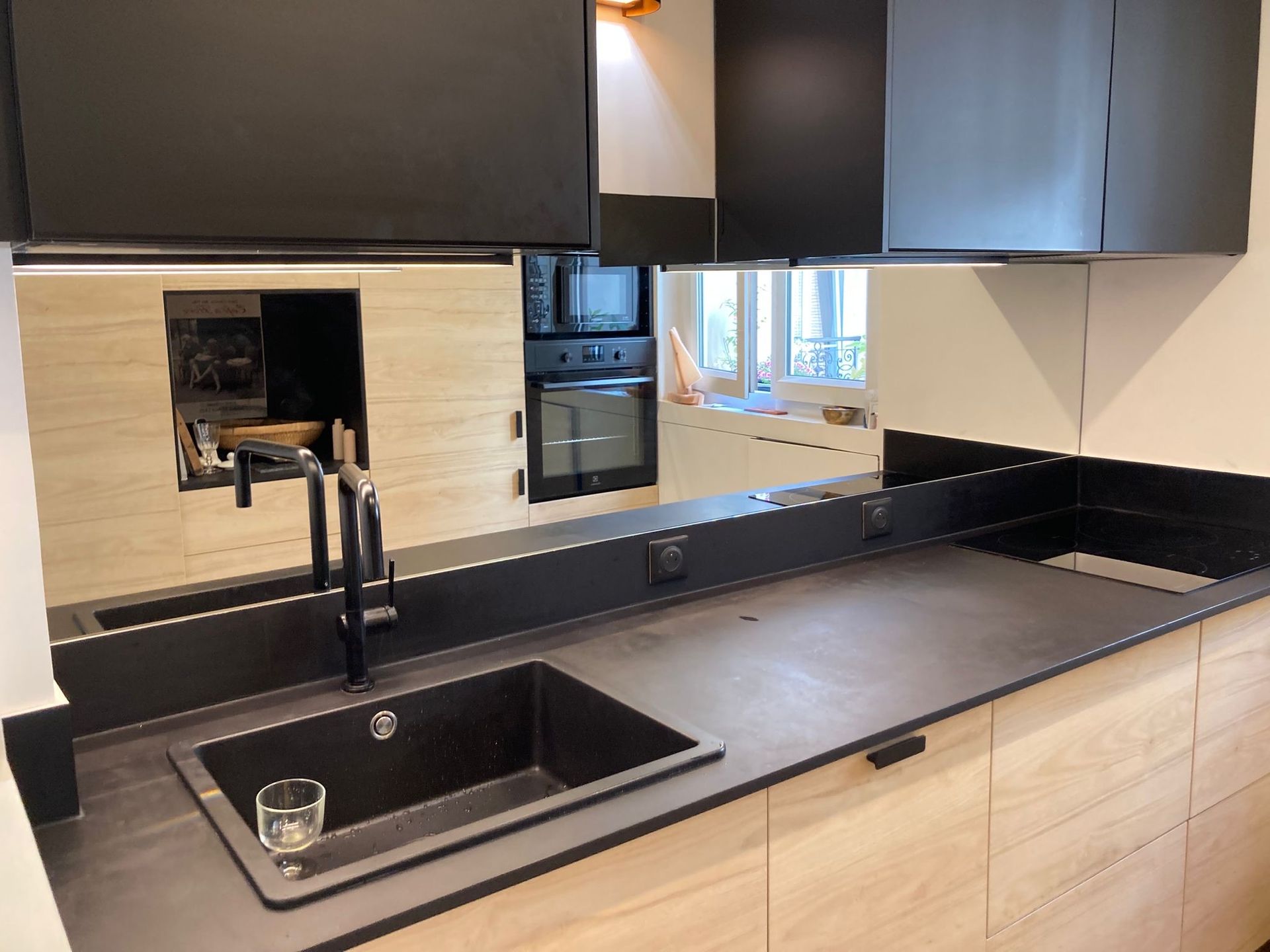 Cuisine moderne avec comptoirs, évier et armoires noirs, un grand miroir en crédence et des armoires de couleur bois clair.