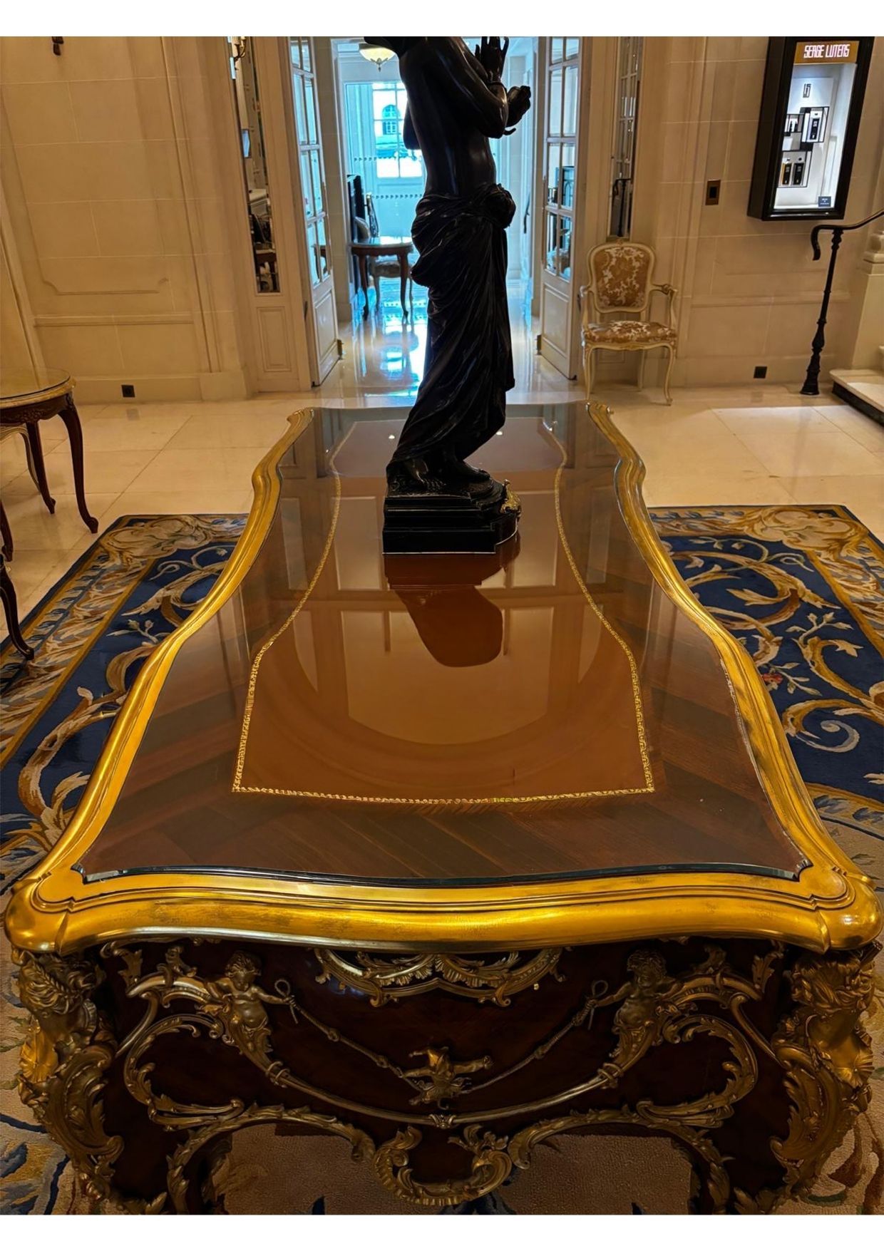 Table en bois ornée de dorures, surmontée d'une sculpture, posée sur un tapis à motifs dans une pièce de réception.