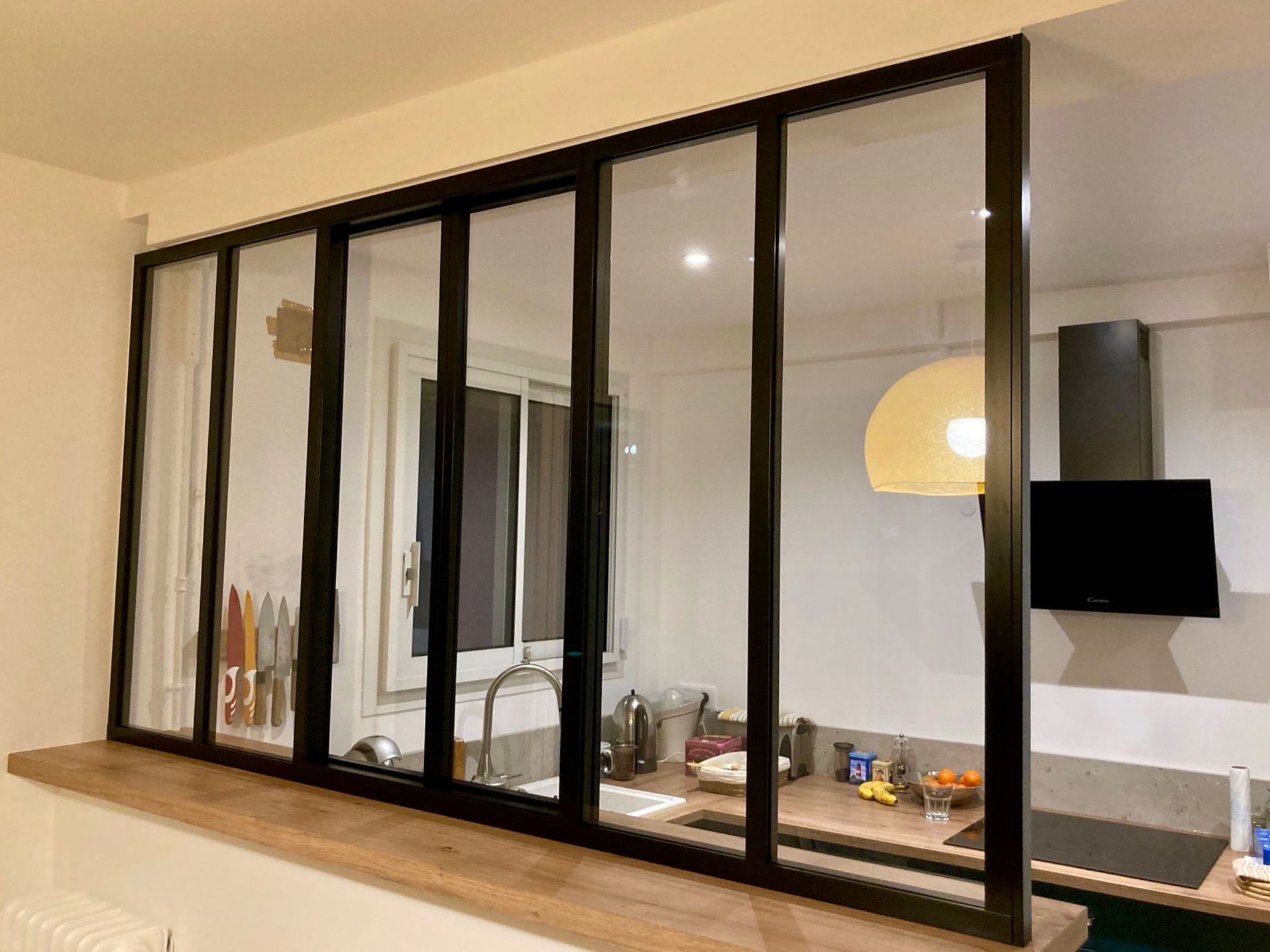 Cloison vitrée à cadre noir séparant une pièce d'une cuisine, avec un plan de travail en bois.