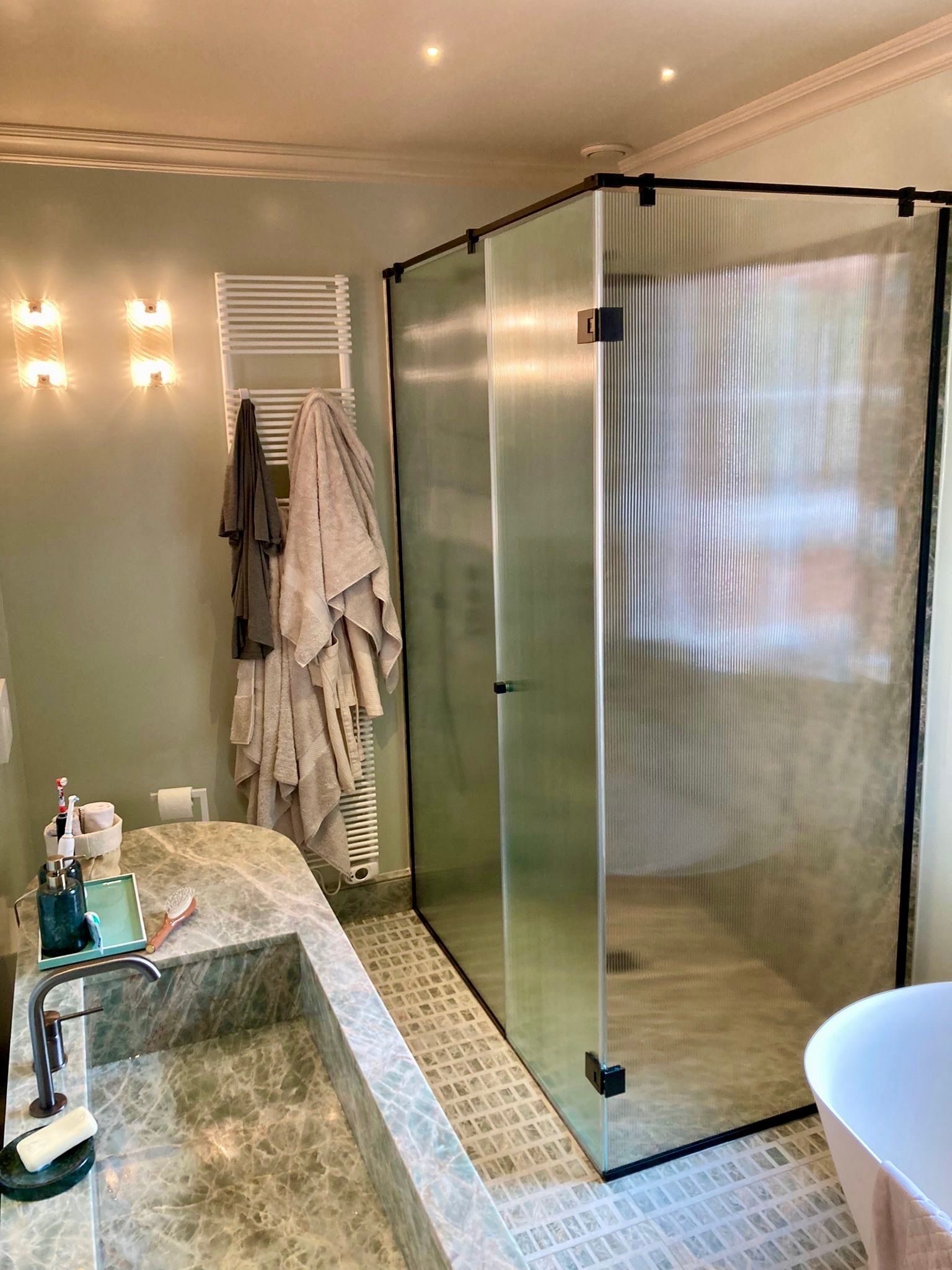 Salle de bains avec douche à l'italienne, baignoire et sèche-serviettes chauffant. Murs vert clair, surfaces en pierre et carrelage.