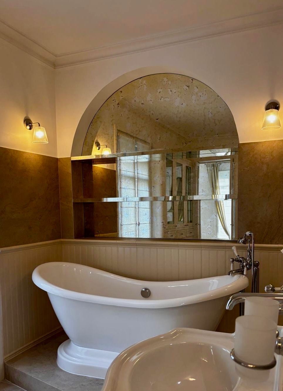 Salle de bains avec baignoire blanche sur pieds, miroir arqué, appliques murales et accents muraux dorés.
