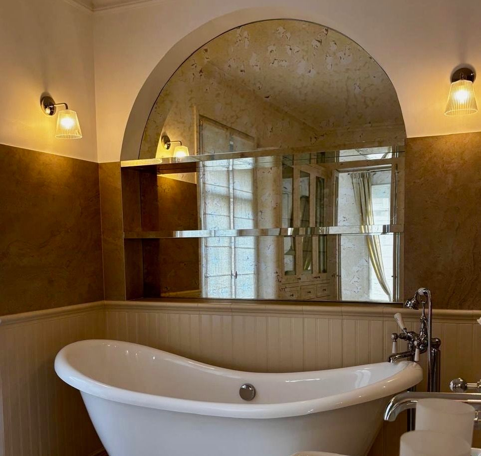 Salle de bains avec baignoire sur pieds, grand miroir arqué, appliques murales et murs aux tons neutres.