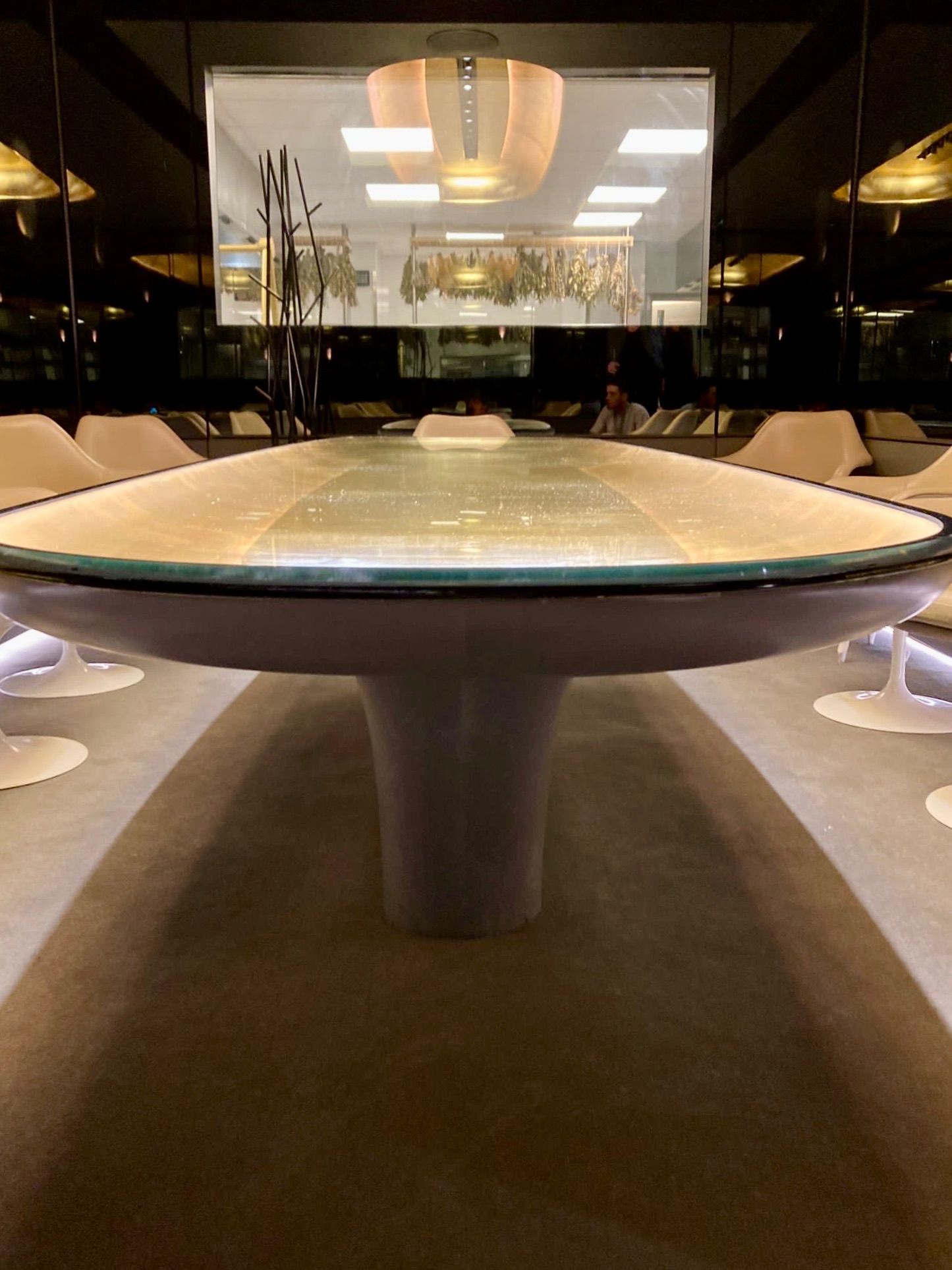 Grande table ovale dans une pièce faiblement éclairée, probablement une salle de conférence. La table possède un pied central et un plateau en verre.