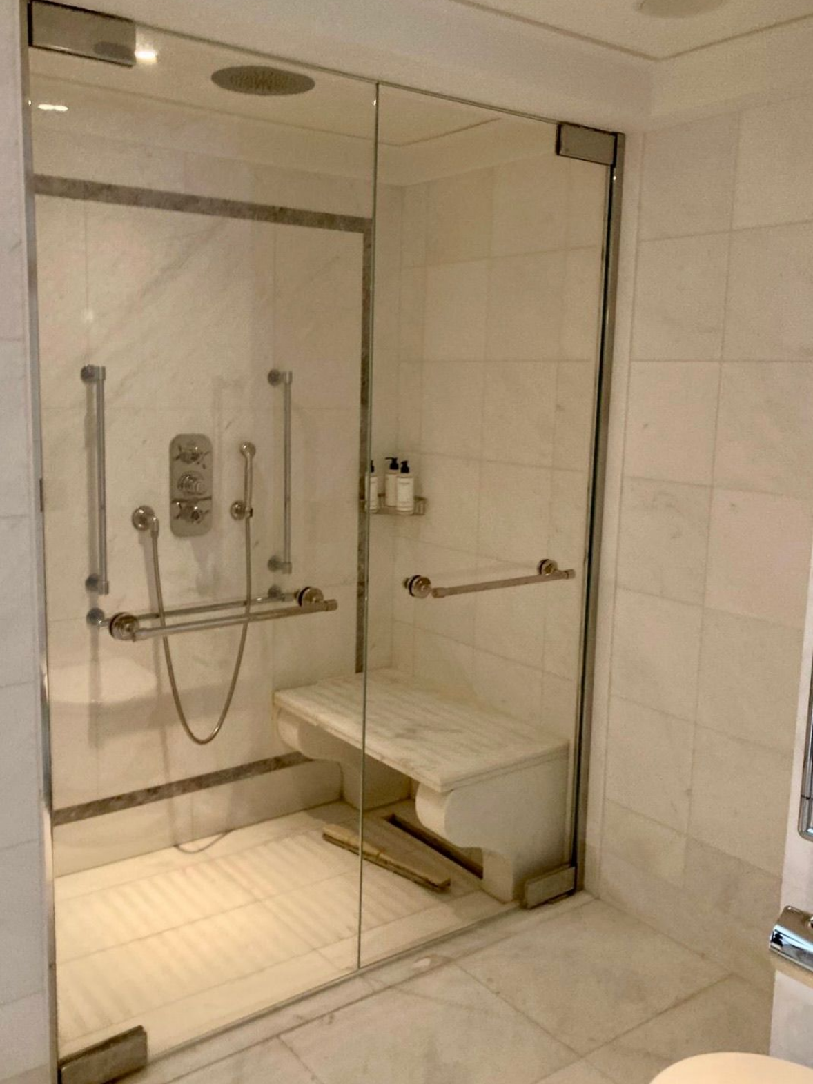Douche accessible carrelée en marbre avec banc, barres d'appui et portes en verre.
