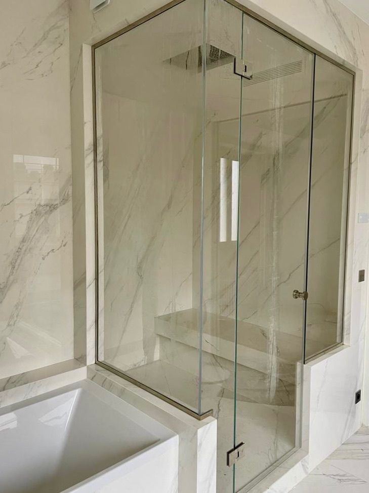 Une cabine de douche en verre sans cadre, avec des murs en marbre blanc et une banquette intégrée, jouxtant une baignoire blanche.