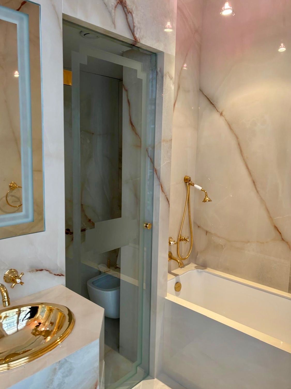 Salle de bains luxueuse avec robinetterie dorée, murs en marbre et douche vitrée jouxtant une baignoire blanche.