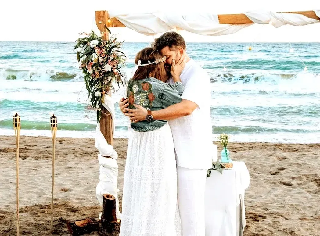 Una novia y un novio se besan bajo un arco de madera en la playa.