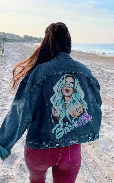 Una mujer que lleva una chaqueta vaquera con una pintura de una mujer en la espalda camina por la playa.
