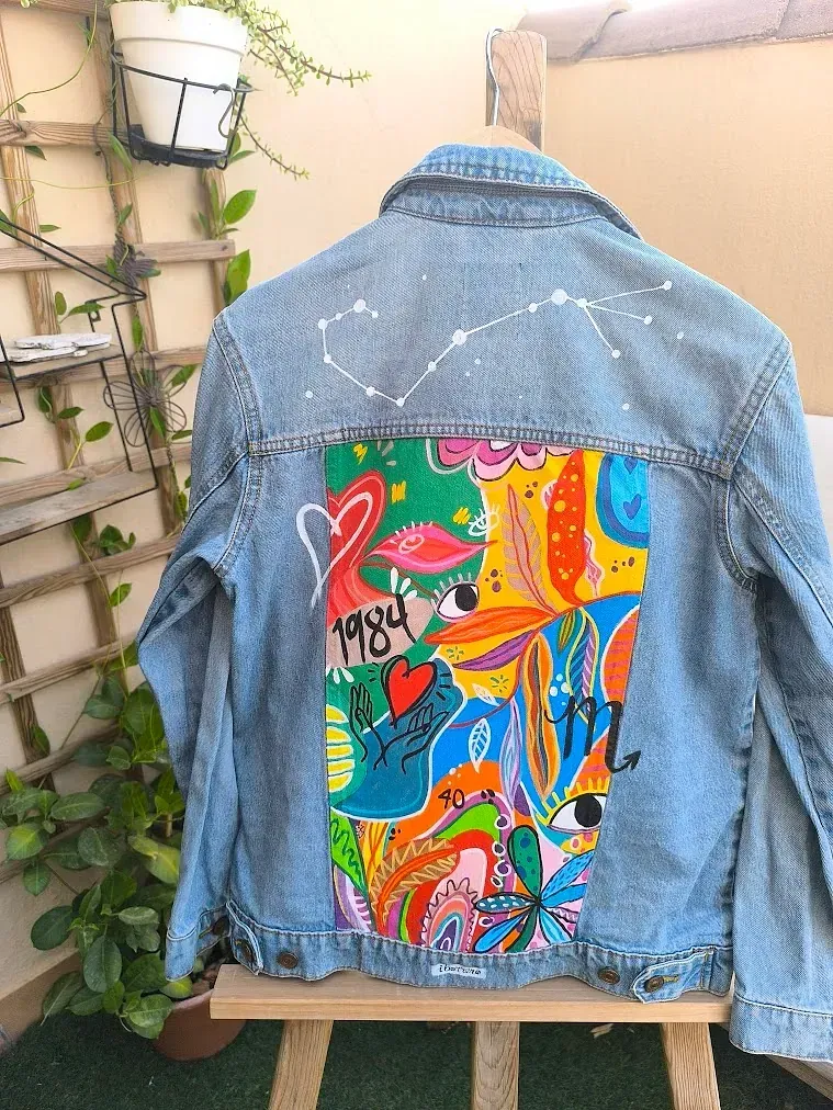 Una chaqueta vaquera con una pintura colorida en la espalda está colocada sobre un caballete de madera.