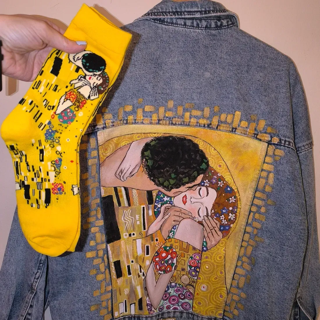 Una persona sostiene un par de calcetines amarillos sobre una chaqueta vaquera con una pintura de un beso en la espalda.
