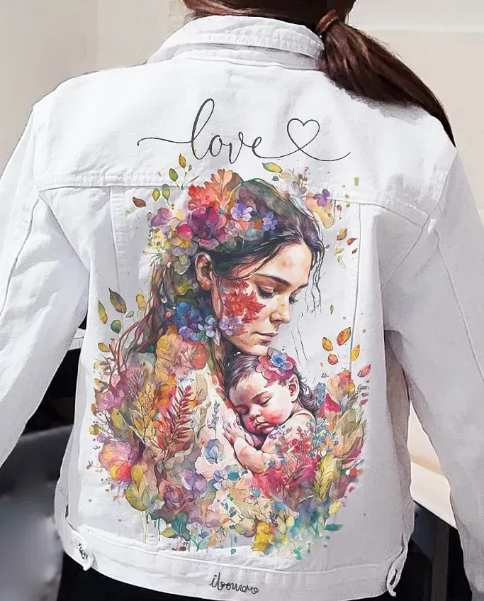 Una mujer lleva una chaqueta vaquera blanca con una pintura de una mujer sosteniendo un bebé en la espalda.