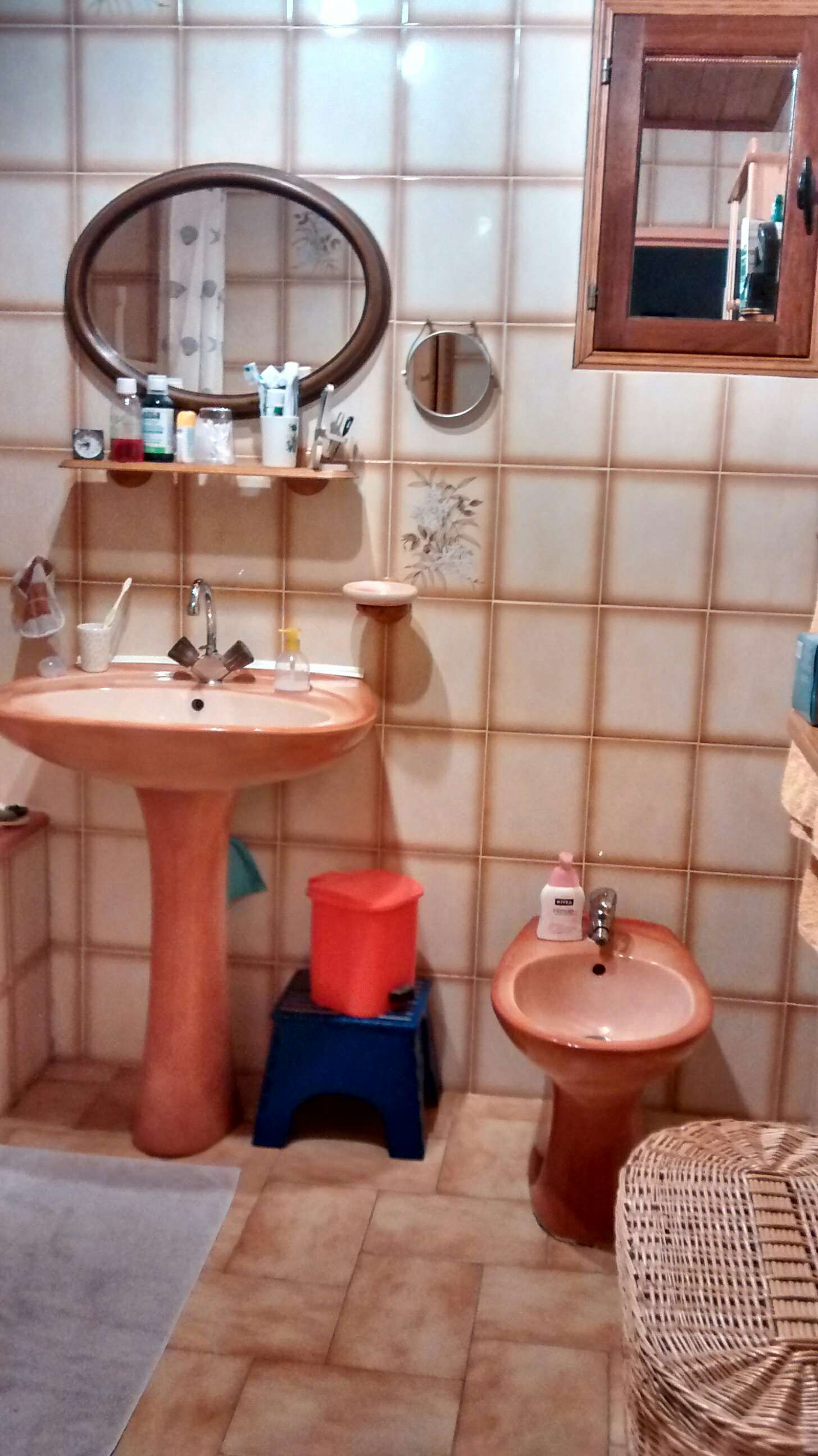 Petite salle de bains carrelée avec lavabo, miroir rond, bidet, toilettes et tabouret bleu avec seau rouge.