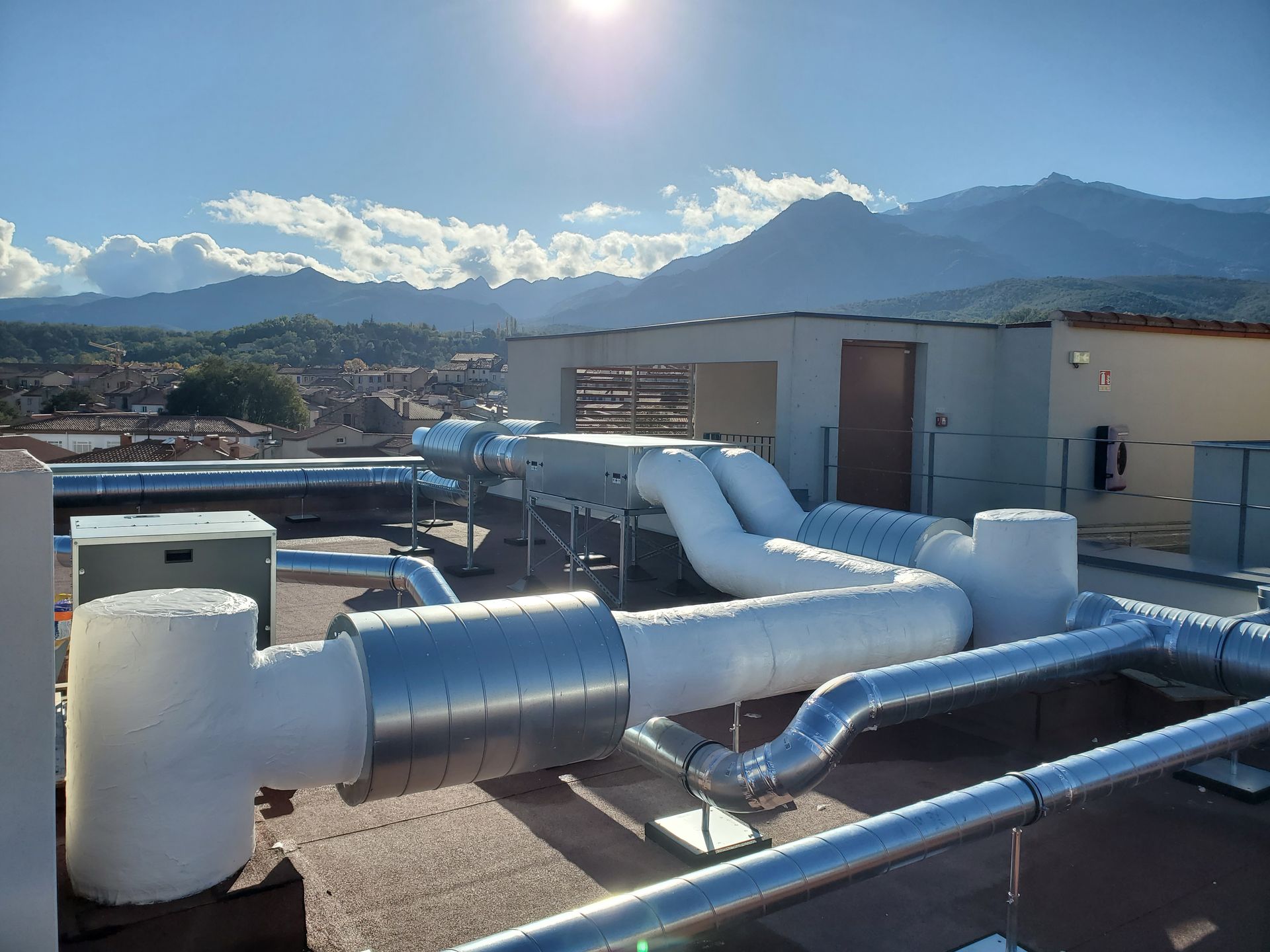 Conduits et bouches d'aération du système de chauffage, ventilation et climatisation sur le toit, sous un soleil éclatant, avec des montagnes et des nuages à l'horizon.