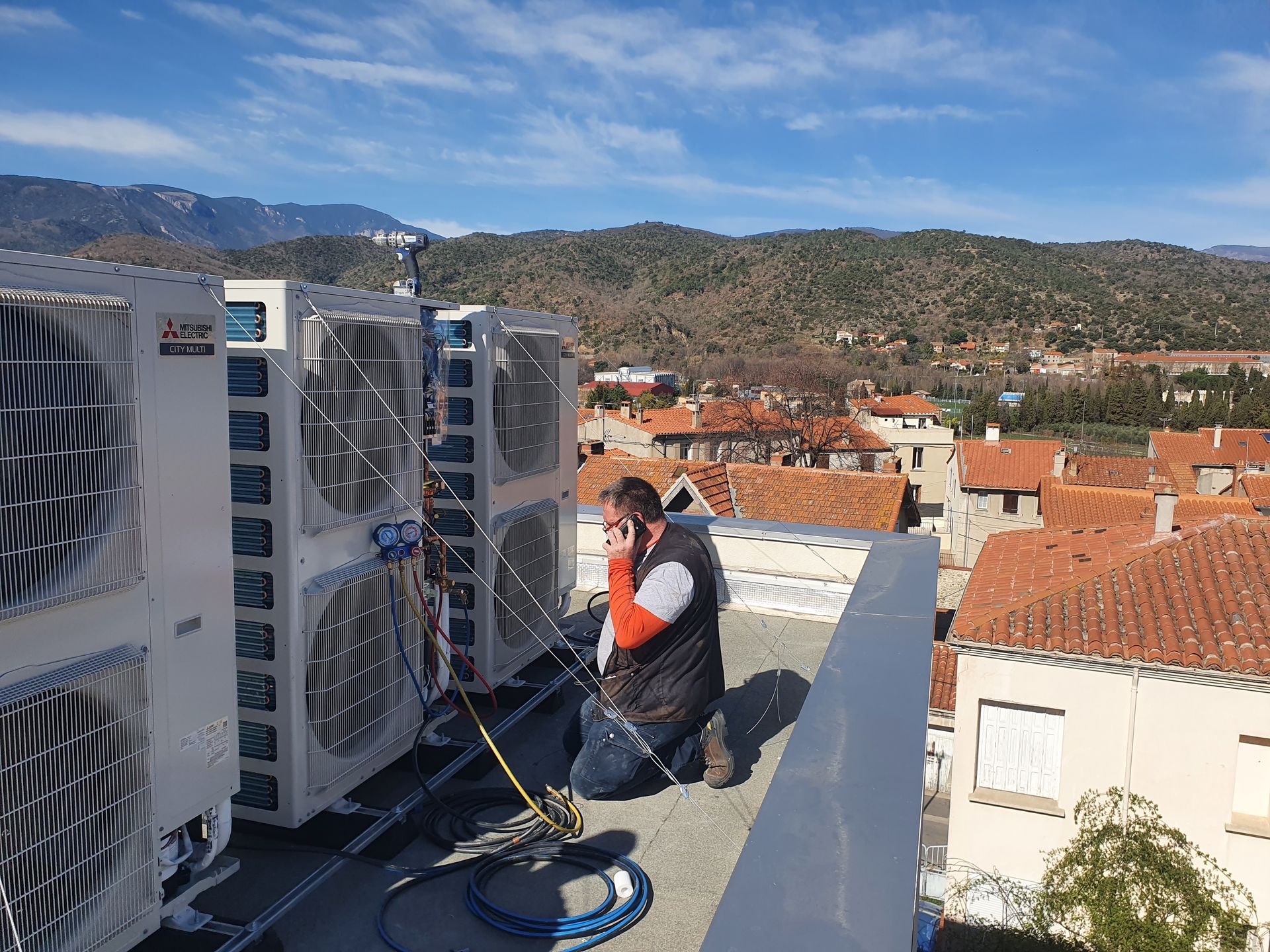 Un technicien travaille à côté d'unités de climatisation sur le toit, avec une ville et des collines en arrière-plan.