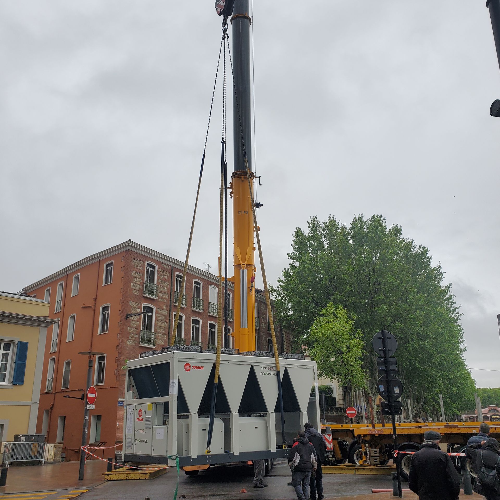 Une grue soulève un grand conteneur industriel blanc à côté d'immeubles en briques dans une rue de la ville.