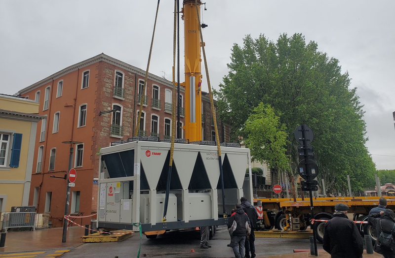 Une grande grue soulève un module industriel blanc à côté d'un bâtiment en briques dans une rue pluvieuse