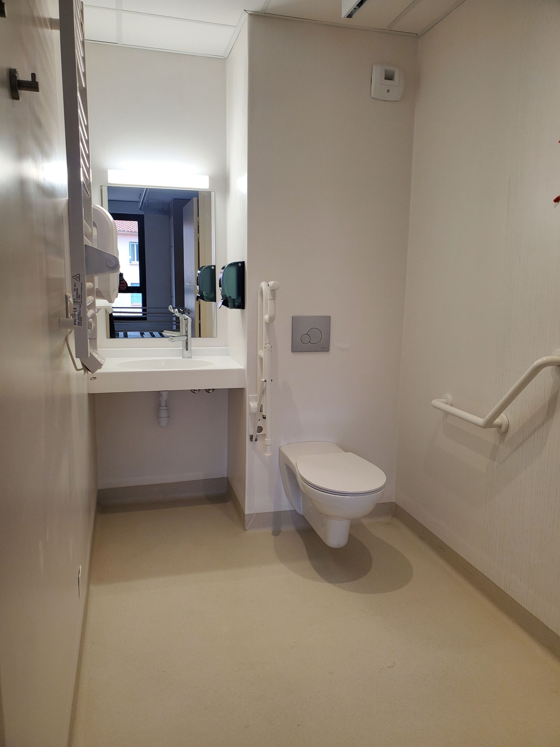 Toilettes accessibles avec W-C, lavabo, barres d'appui et grand miroir dans une pièce beige lumineuse