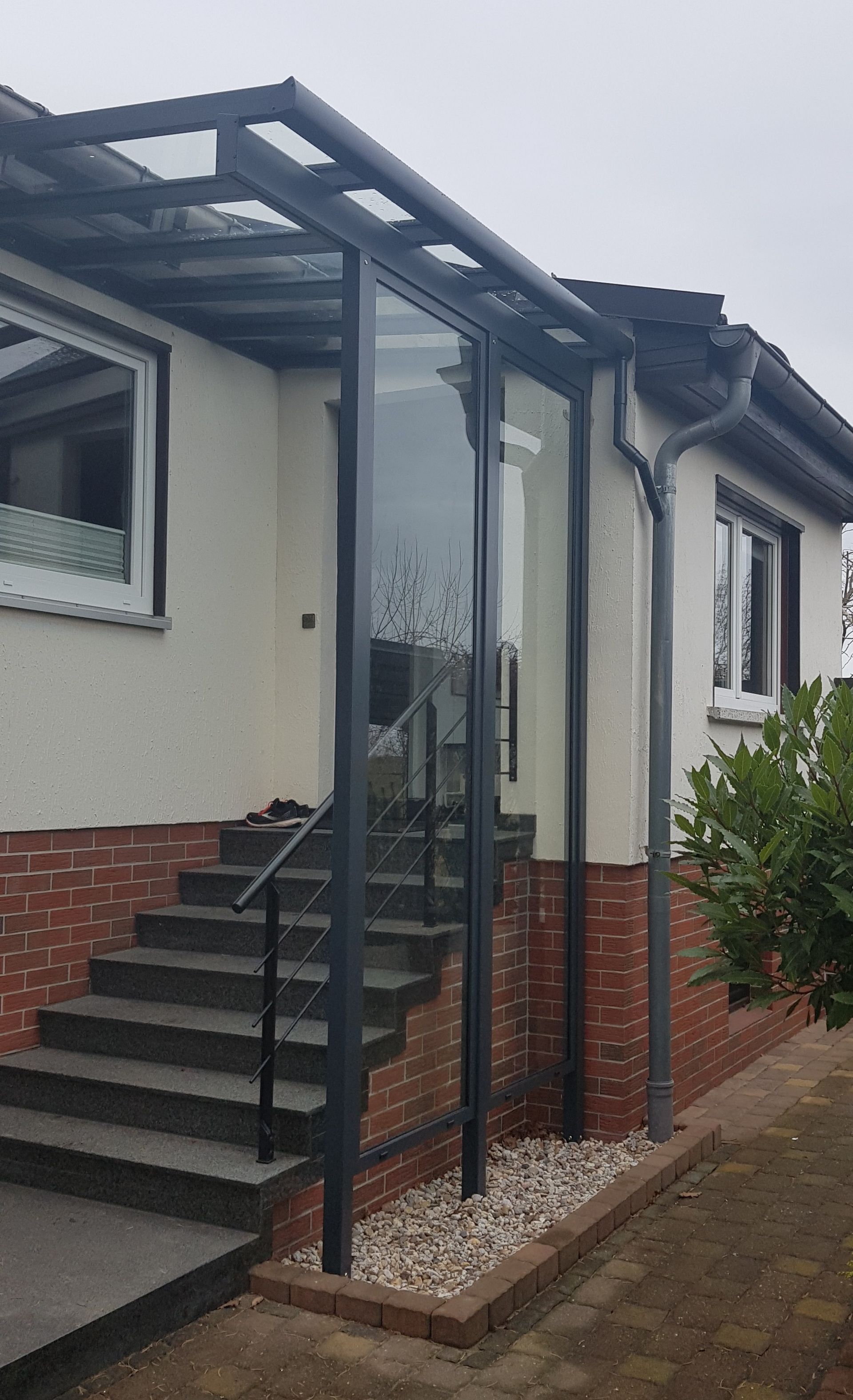 Über einer Außentreppe aus Ziegelsteinen, die zum Hauseingang führt, wurde ein dunkelgraues Metallvordach mit seitlicher Glasscheibe installiert.