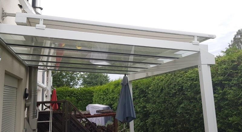 Eine weiße Pergola mit einem klaren Glasdach und einem daran hängenden blauen Sonnenschirm.