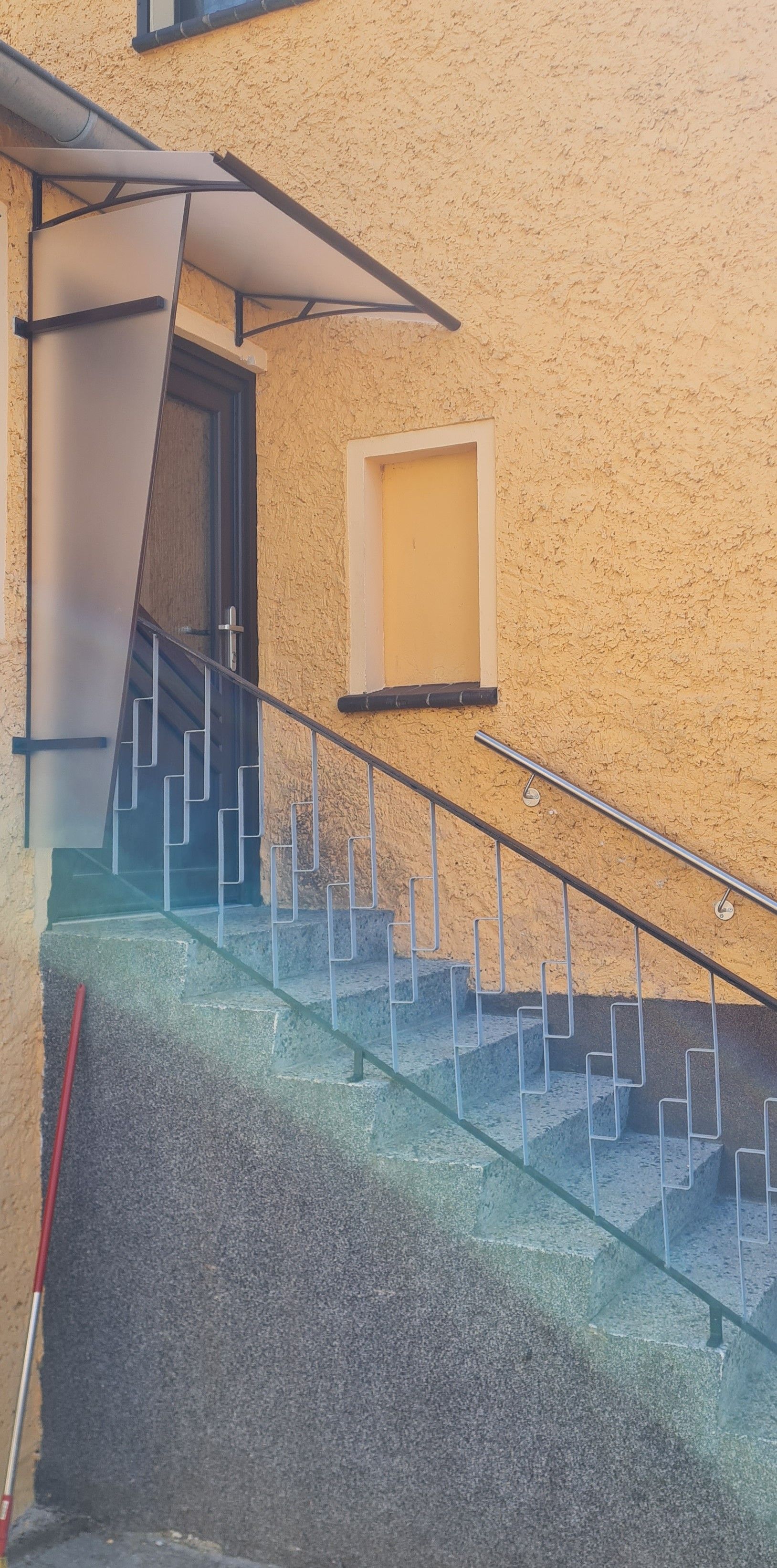 Eine Außentreppe mit Metallgeländer führt zu einer dunklen Tür unter einer kleinen Markise an einer gelben, strukturierten Gebäudewand.