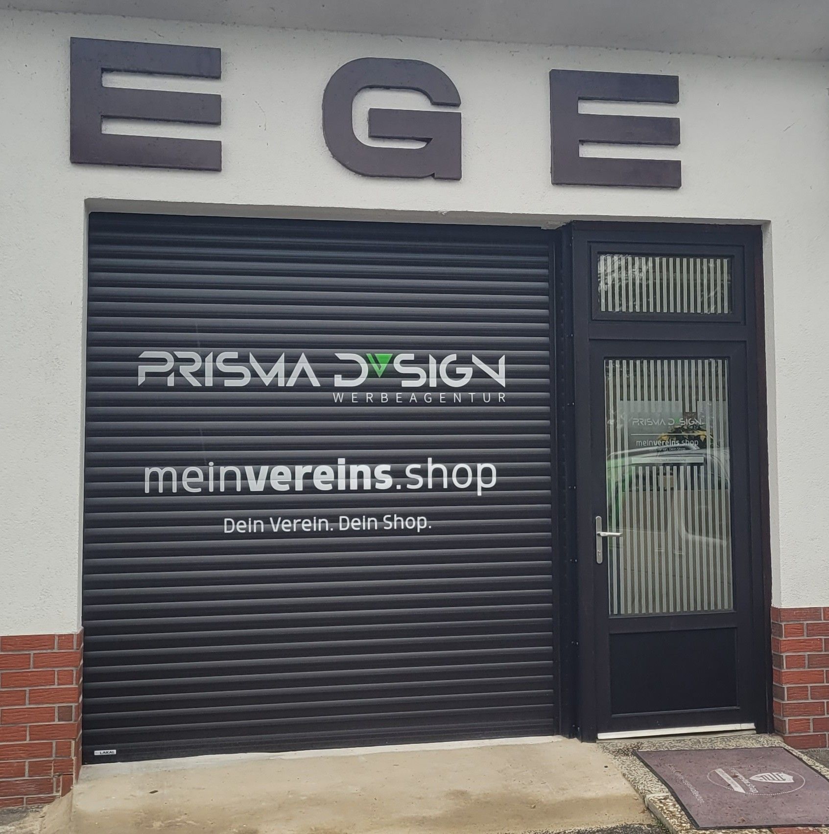 Schaufenster mit „EGE“-Schild, schwarzem Rollladen und Tür. „PRISMA DESIGN“-Logo und „meinvereins.shop“ auf dem Rollladen.