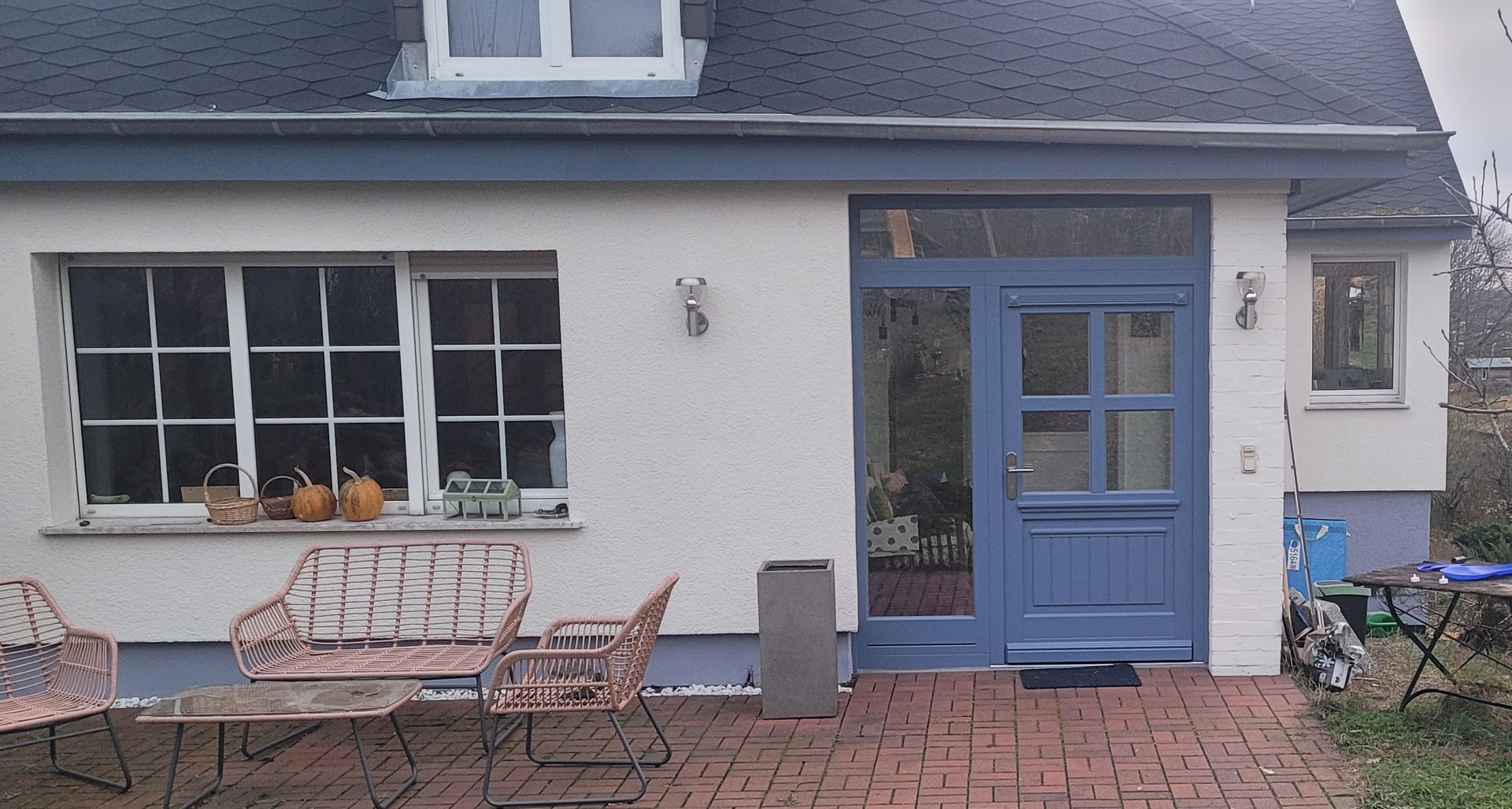 Weißes Haus mit blauer Tür und Zierleisten. Backsteinterrasse, Gartenmöbel aus Metall und Fenster mit Kürbissen.
