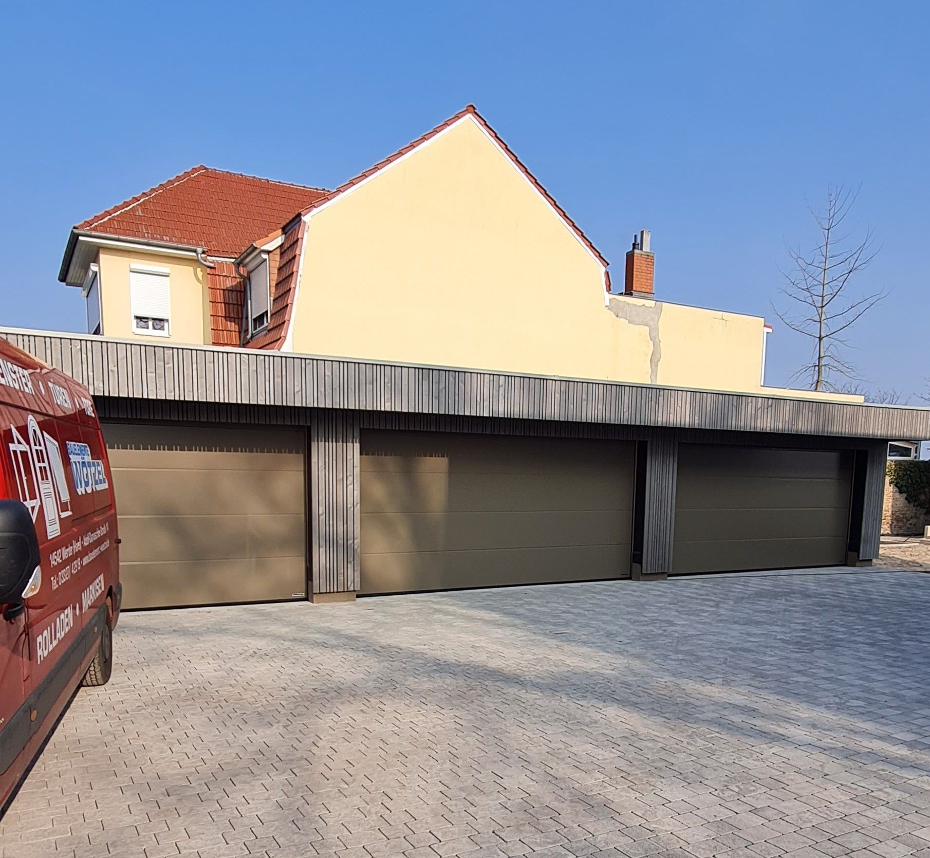 Eine Dreifachgarage mit hellbraunen Toren und einer Fassade aus Holzlatten, vor einem cremefarbenen Haus und einer hellen Backsteinauffahrt.